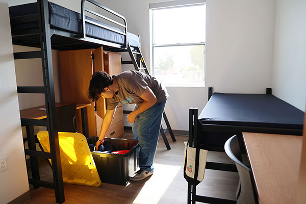 A student organizes belongings in a storage bin inside a furnished room with bunk beds, a single bed, a desk, and a chair.
