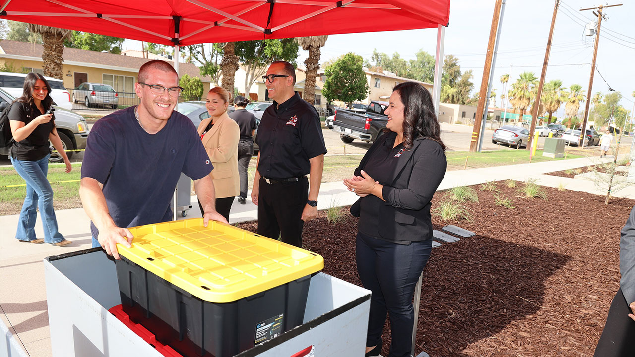College staff members gather under a red canopy as a students moves a large storage container while others look on during an outdoor campus event.
