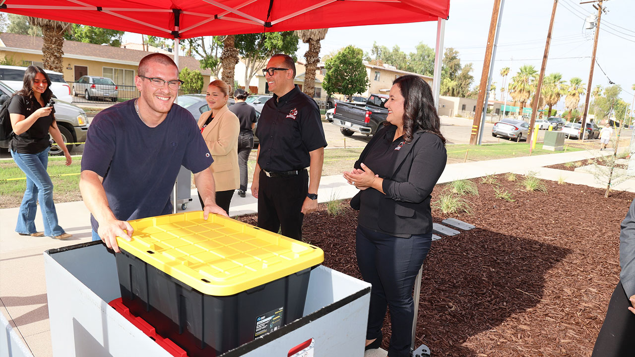 College staff members gather under a red canopy as a students moves a large storage container while others look on during an outdoor campus event.
