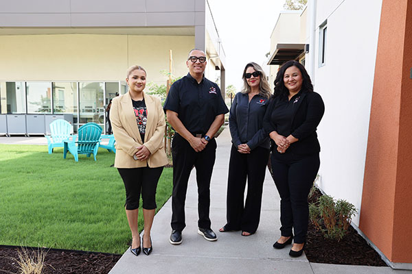 Four college staff members standing together outside a campus building, with a grassy courtyard and colorful outdoor chairs in the background.