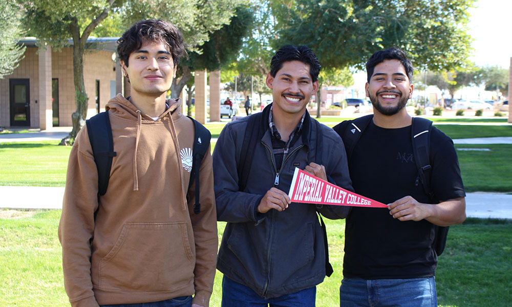 Three Imperial Valley College students stand on campus wearing backpacks, with two holding an Imperial Valley College pennant and smiling for the camera.