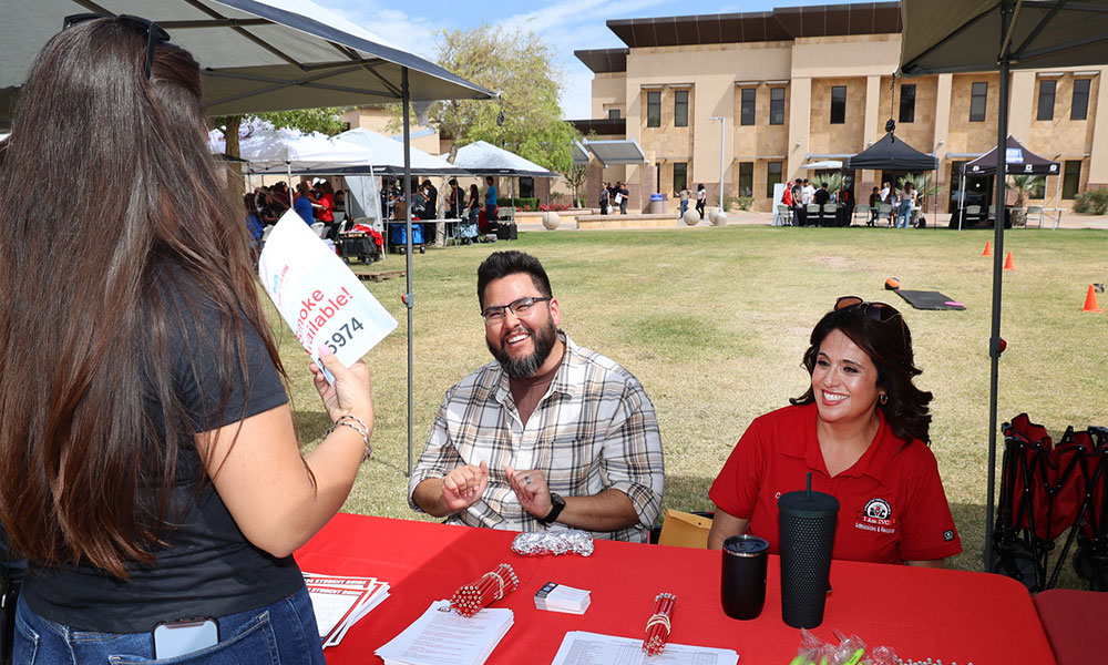 Two Imperial Valley College staff members speak with a visitor at an outdoor information table during a campus event, with additional booths and the college campus in the background.