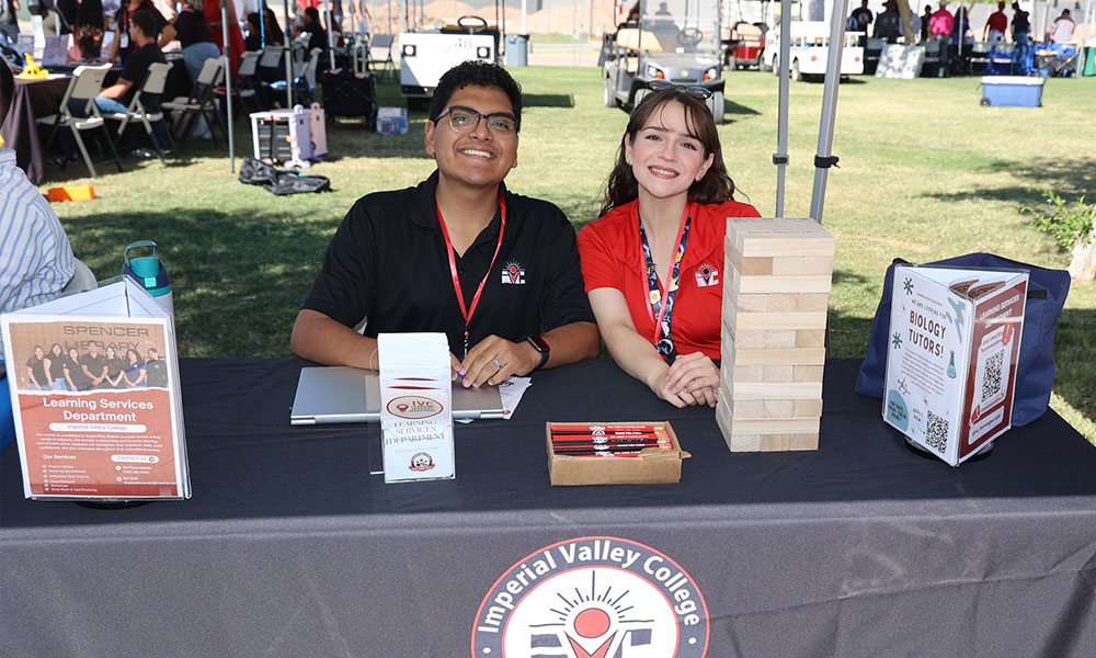Two Imperial Valley College staff members sit at a campus event information table, with promotional materials, a Jenga-style game, and a Biology Tutors display in front of them.