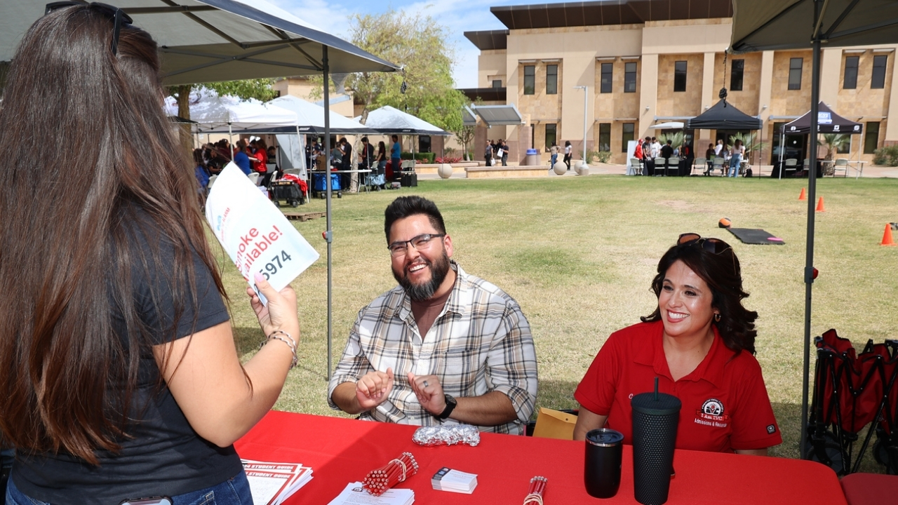 Two Imperial Valley College staff members speak with a visitor at an outdoor information table during a campus event, with additional booths and the college campus in the background.