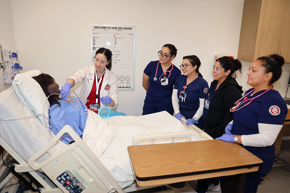 ursing students in scrubs practice patient care around a hospital bed with a simulated patient mannequin in a clinical simulation center.